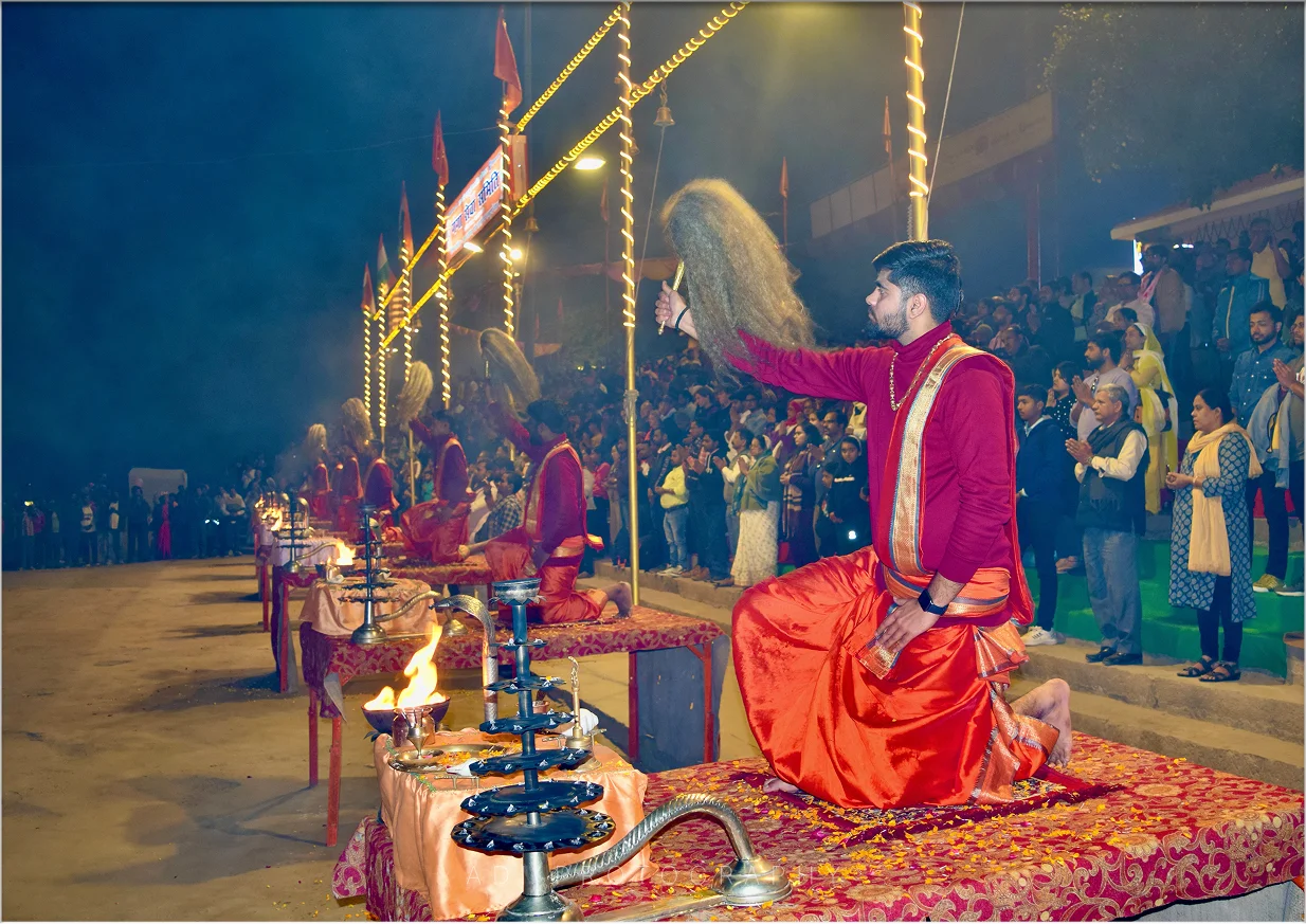 Photograph of a kneeling priest raising a chamar at the Ganga Aarti in Varanasi with lamps and crowd behind